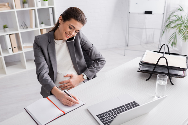 χαρούμενη έγκυος επιχειρηματίας μιλώντας στο smartphone και γράφοντας στο σημειωματάριο κοντά στο φορητό υπολογιστή στο γραφείο - Φωτογραφία, εικόνα
