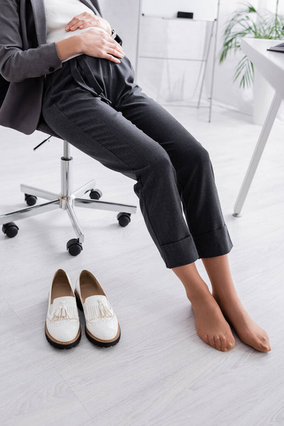 vista recortada de mujer embarazada sin zapatos sentada en la silla cerca del escritorio - Foto, Imagen