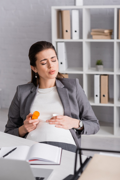 mulher grávida segurando jar com pílulas enquanto se sente cãibra no escritório - Foto, Imagem