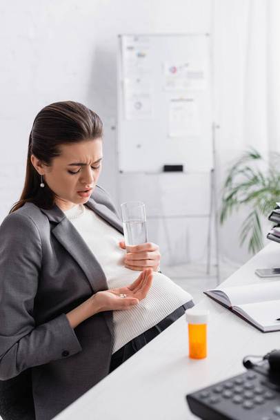 pregnant woman holding pills and glass of water while feeling cramp in office - Foto, afbeelding