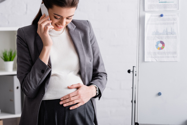 joyful pregnant businesswoman talking on smartphone near flip chart in office - Фото, изображение