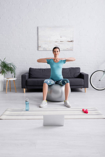 happy pregnant woman in sportswear exercising on fitness ball near laptop in living room  - Foto, imagen