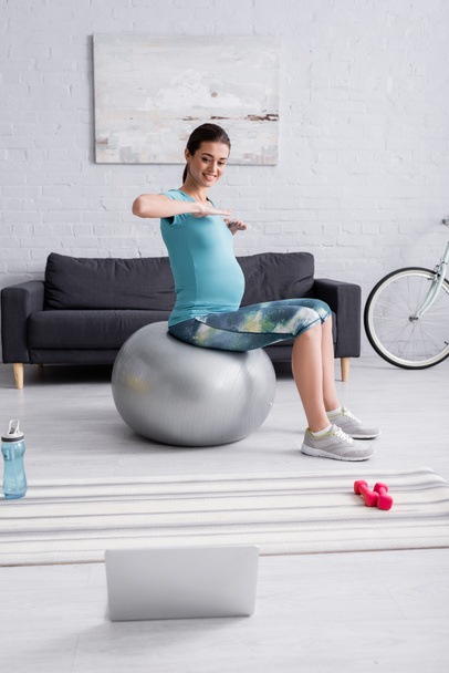Spor giyimli hamile bir kadın oturma odasında dizüstü bilgisayarın yanında spor topuyla egzersiz yapıyor.  - Fotoğraf, Görsel