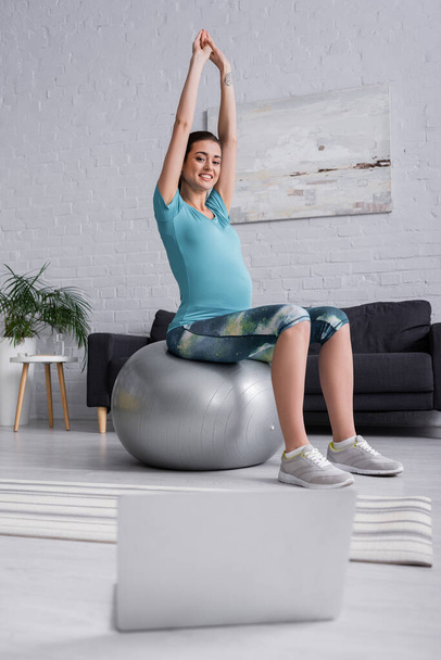 pozitivní těhotná žena se zvednutýma rukama cvičení na fitness míč a při pohledu na notebook v obývacím pokoji  - Fotografie, Obrázek