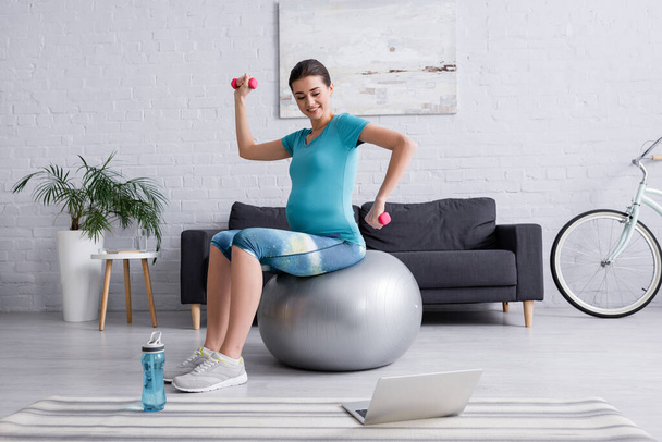 allegra donna incinta che si esercita sulla palla fitness con manubri vicino al computer portatile - Foto, immagini