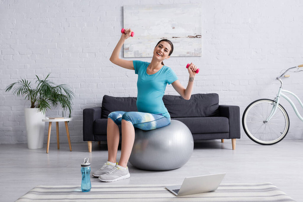 счастливая беременная женщина упражняется на фитнес-мяч с гантелями возле ноутбука - Фото, изображение