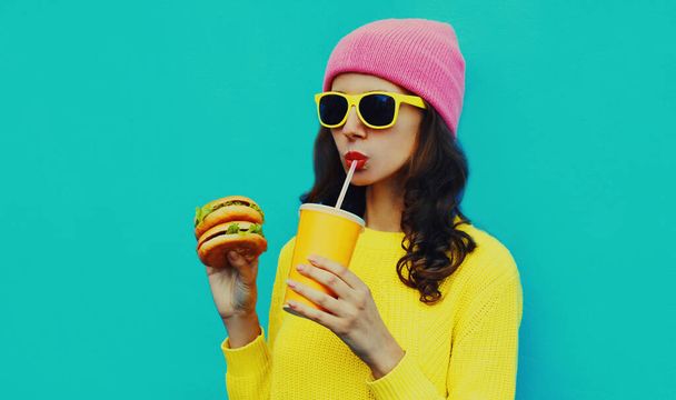 Portrait of stylish young woman drinking juice with burger fast food on blue colorful background - Foto, Imagem