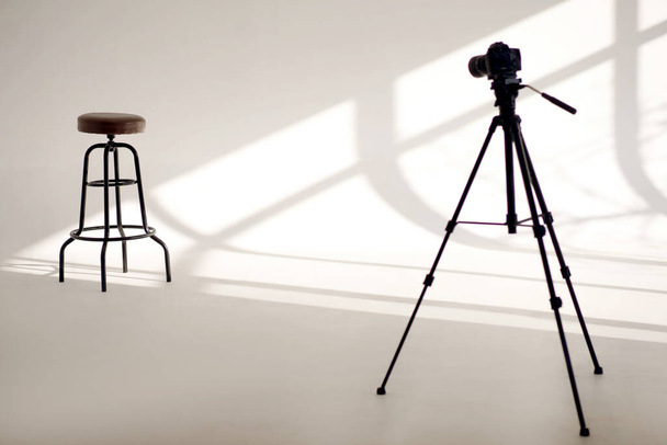 Un estudio fotográfico blanco con sombras de sol desde las ventanas y una silla alta y una cámara de video en un trípode. Fotografía profesional con taburete de bar para modelo y espacio en blanco con sombras - Foto, imagen