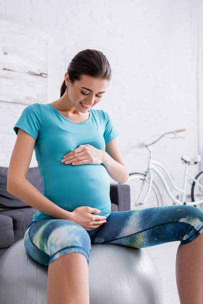 cheerful pregnant sportswoman sitting on fitness ball and touching belly - Fotoğraf, Görsel