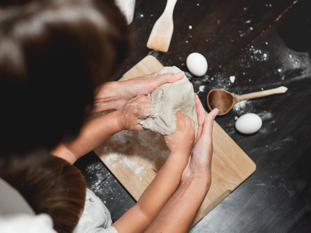 Mother and son are cooking together. Mom and toddler knead dough from flour and eggs on black wooden table. Family time. Fun at kitchen. Developing fine motor skills. - Valokuva, kuva