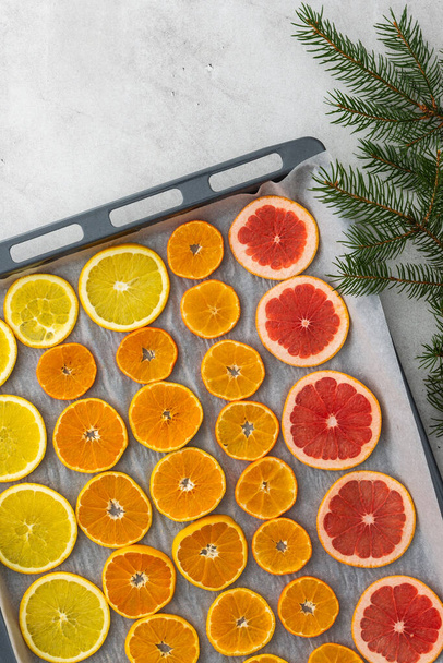 φέτες πορτοκαλιού, μανταρινιού και γκρέιπφρουτ σε ταψί και κλαδιά ελάτης, ετοιμάζοντας φέτες εσπεριδοειδών για Χριστουγεννιάτικες γιρλάντες - Φωτογραφία, εικόνα