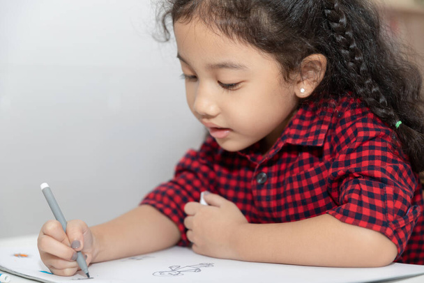 Малышка азиатка в клетчатой рубашке сидит и рисует на бумажной книге на белом столе в комнате - Фото, изображение