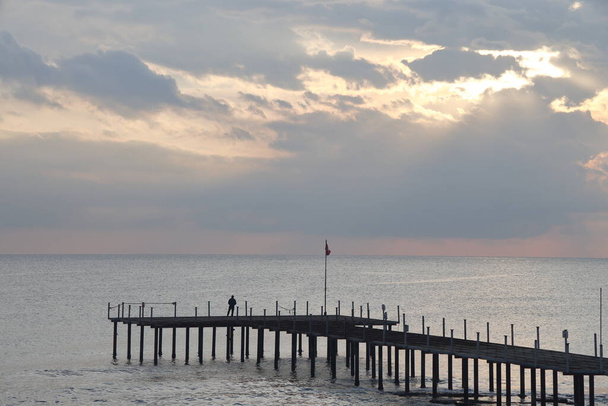 Silhouettes on the pier against the backdrop of the calm sea and the sun making its way through the clouds. - Fotó, kép