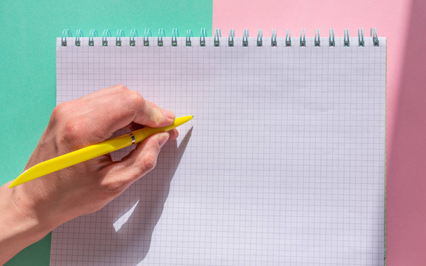 The man is holding a yellow pen with his left hand and is about to write on a pad of paper. Left-handed day. Southpaw. Pink and turquoise background. - Φωτογραφία, εικόνα