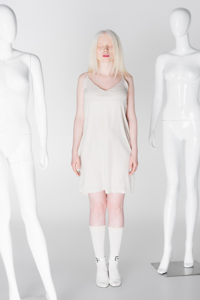 Volledige lengte van albino model in jurk staan in de buurt van etalagepoppen op witte achtergrond  - Foto, afbeelding