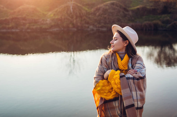 Young woman relaxing by autumn lake at sunset. Stylish girl in hat enjoying fall landscape, breathing freely. Mental health. Healing nature - Photo, image