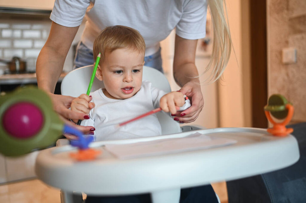 Kicsi aranyos kisbaba szőke fiú ül babakocsin, és rajzolni tanul. Gyönyörű fiatal anya és fia játszanak időt együtt otthon játékokkal. Egészséges boldog családi gyermekkori koncepció. - Fotó, kép