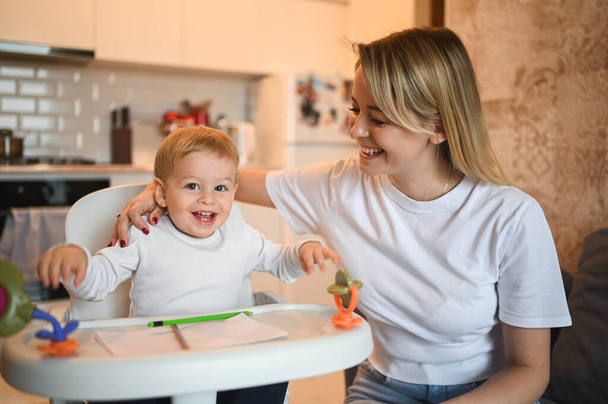 Little cute baby toddler boy blonde sitting on baby chair learning to draw. Beautiful young mom and son playing spend time together indoors at home with toys. Healthy happy family childhood concept. - Photo, Image
