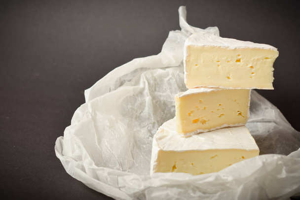 Κοντινό πλάνο από μαλακά κομμάτια γαλλικού τυριού σε λευκό περγαμηνό χαρτί σε σκούρο φόντο. Τρία κομμάτια τυρί. - Φωτογραφία, εικόνα