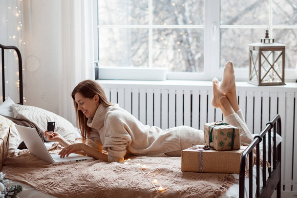 Woman is laying on the bed and shopping online using her laptop and credit card, Christmas time.  - Fotoğraf, Görsel
