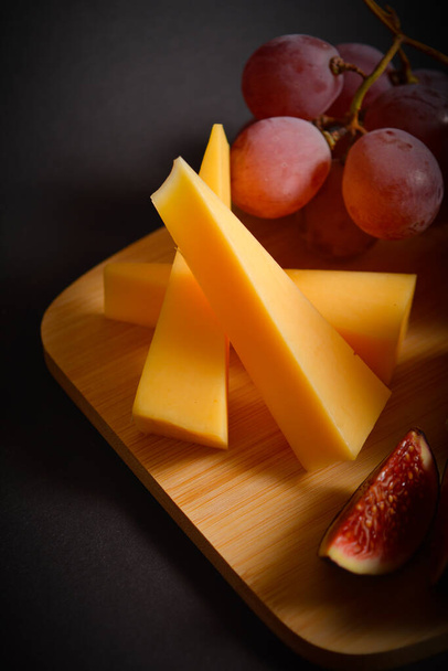 Osa juusto aluksella kovaa juustoa, punaisia viinirypäleitä ja viikunan paloja. Lautanen mustalla taustalla. Pystysuora kuva. Alkupaloja ja alkupaloja. Tumma tunnelma valokuva tyyli - Valokuva, kuva
