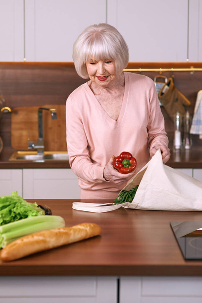 Старшая веселая женщина переупаковывает продукты на современной кухне. Еда, навыки, образ жизни - Фото, изображение