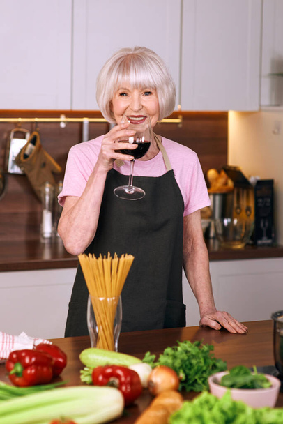 ηλικιωμένη χαρούμενη γυναίκα πίνει κόκκινο κρασί κατά τη διάρκεια του μαγειρέματος στη σύγχρονη κουζίνα. Τρόφιμα, εκπαίδευση, τρόπος ζωής - Φωτογραφία, εικόνα