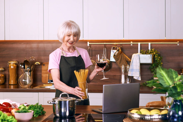 ηλικιωμένη χαρούμενη γυναίκα μαγειρεύει στη σύγχρονη κουζίνα με φορητό υπολογιστή. Τρόφιμα, εκπαίδευση, τρόπος ζωής  - Φωτογραφία, εικόνα
