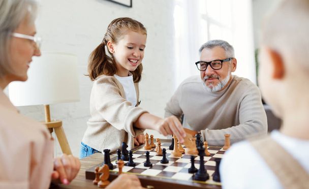 Дети брат и сестра играют в шахматы, сидя в гостиной со старшими бабушкой и дедушкой - Фото, изображение