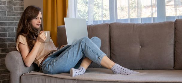 Ένα πανό μιας νεαρής γυναίκας πίνει ένα φλιτζάνι καφέ κατά τη διάρκεια της εργασίας από το σπίτι. Ένα κορίτσι σκέφτεται και κάθεται στον καναπέ με ένα φορητό υπολογιστή - Φωτογραφία, εικόνα