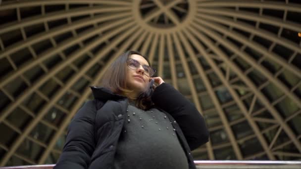 Μια νεαρή έγκυος με γυαλιά και μπουφάν στέκεται και μιλάει στο τηλέφωνο. Ένα κορίτσι σε ένα εμπορικό κέντρο χρησιμοποιεί ένα smartphone με φόντο ένα πανοραμικό θόλο. - Πλάνα, βίντεο