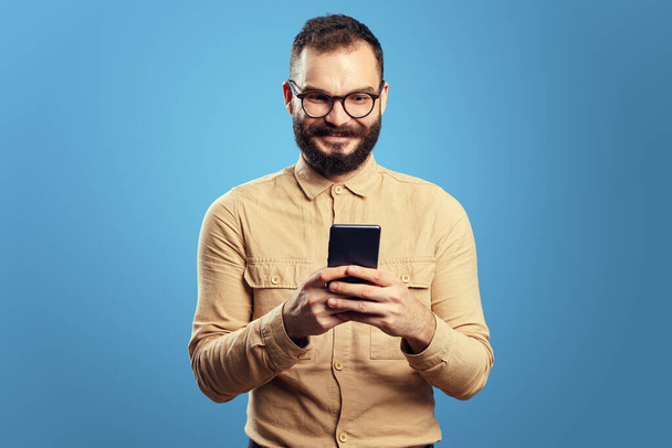 Мужчина улыбается и использует смартфон, стоя изолированно на синем фоне - Фото, изображение