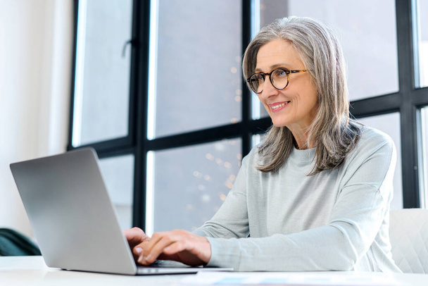 Cheerful business lady wearing glasses typing on the keyboard, looks at the screen satisfied - Foto, Bild