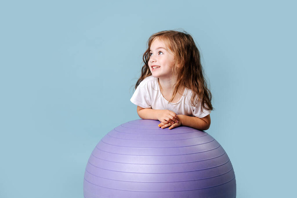 blue-eyed curly babe with dimples on her cheeks laughs plays on the lilac fitness ball - Zdjęcie, obraz