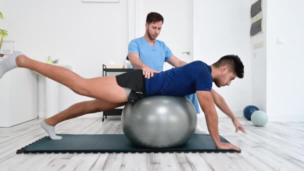 Fysiotherapeut helpt knappe jonge patiënt voor het doen pilates oefeningen met bal. - Video