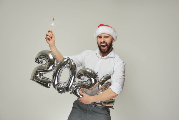 Kırmızı kadife Noel Baba şapkalı gülümseyen bir adam bir elinde 2022 şeklinde gümüş balonlar diğer elinde ise maytap tutuyor. Sakallı bir adam yeni yıl partisinde bağırıyor.. - Fotoğraf, Görsel