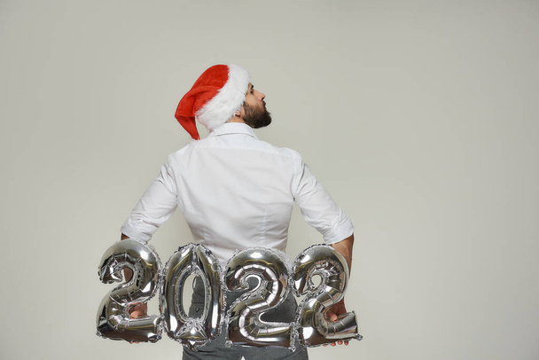 Человек в красной бархатной шляпе Санта поднимает за спиной серебряные шарики в форме 2022 года. Парень с бородой повернул голову направо на новогодней вечеринке.. - Фото, изображение