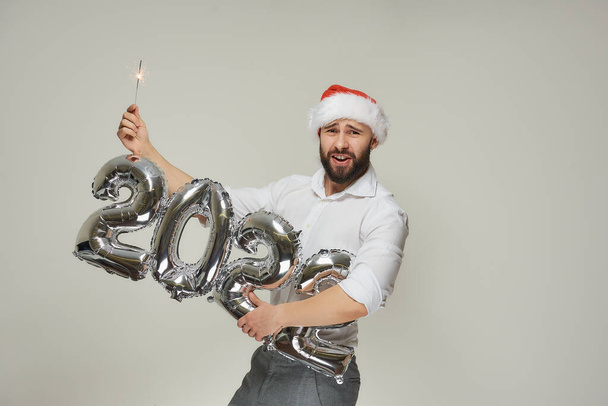 Чоловік у червоному оксамитовому капелюсі Санта тримає однією рукою срібні кульки у формі 2022 року та блискавку іншою рукою. Щасливий хлопець з бородою здивований на новорічній вечірці
. - Фото, зображення