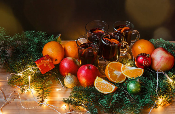 Glazen glaasjes glühwein, appels, sinaasappels, kerstballen, cadeau, dennentakken en gloeiende kerstverlichting op houten tafel. Selectieve focus. - Foto, afbeelding