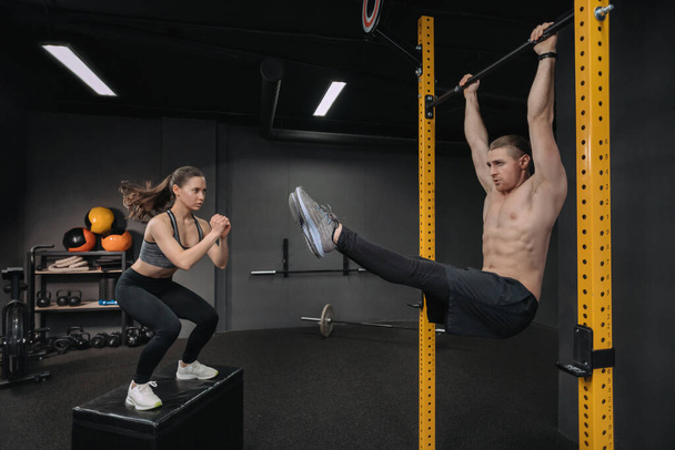 Trening crossfit. Przystojny, muskularny mężczyzna robi ćwiczenia brzucha, podczas gdy wysportowana kobieta wykonuje ćwiczenia skokowe. Trening funkcjonalny, trening grupowy, zajęcia fitness - Zdjęcie, obraz