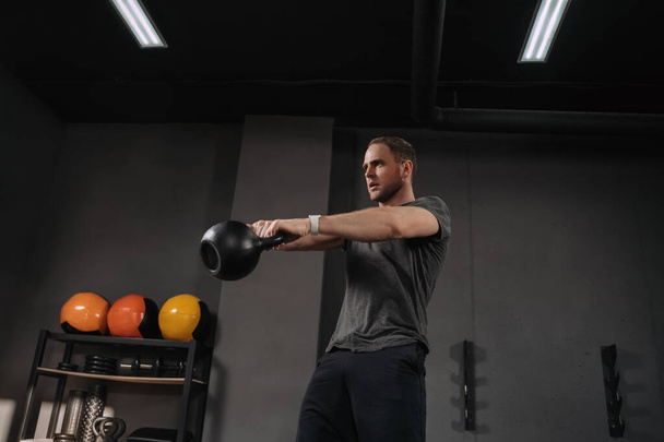 Strong muscular athlete doing kettlebell swings at crossfit gym, outfit shot on grey gym background with equipment. Weightlifter doing workout indoors, exercising. Crossfit instructor warming up - Photo, Image