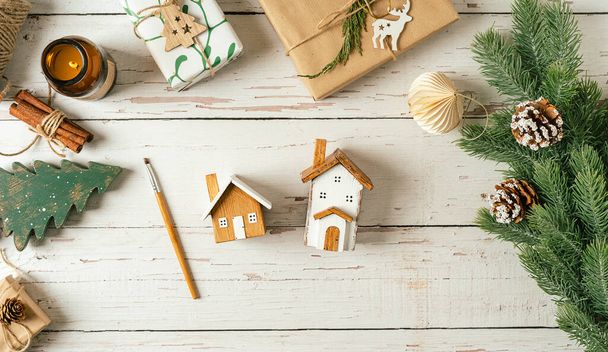 Flat lay of white wooden background with Christmas natural eco decoration. Homemade wooden house with painting brush. Top view gifts, candle, fir tree, toy, cinnamon, pine cone, paper with copy space - Photo, image