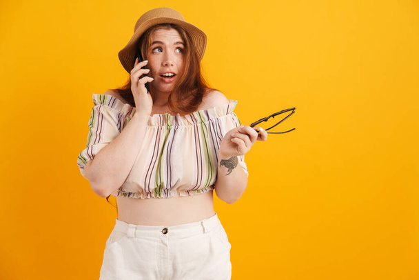 Young ginger woman in hat talking on mobile phone isolated over yellow background - Photo, Image