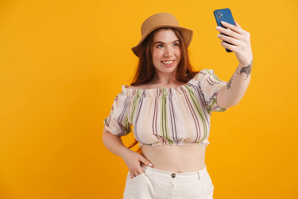 Young ginger woman in hat taking selfie on mobile phone isolated over yellow background - Zdjęcie, obraz