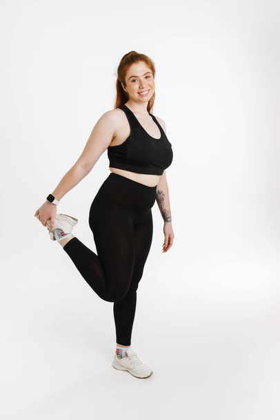 Smiling young plus size fitness woman in sportswear standing over white background doing stretching exercises - Foto, afbeelding