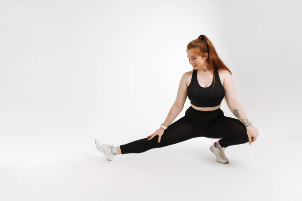 Smiling young plus size fitness woman in sportswear standing over white background doing stretching exercises - Foto, immagini