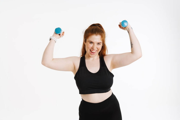 Smiling young plus size fitness woman in sportswear standing over white background exercises with dumbbells - Fotografie, Obrázek