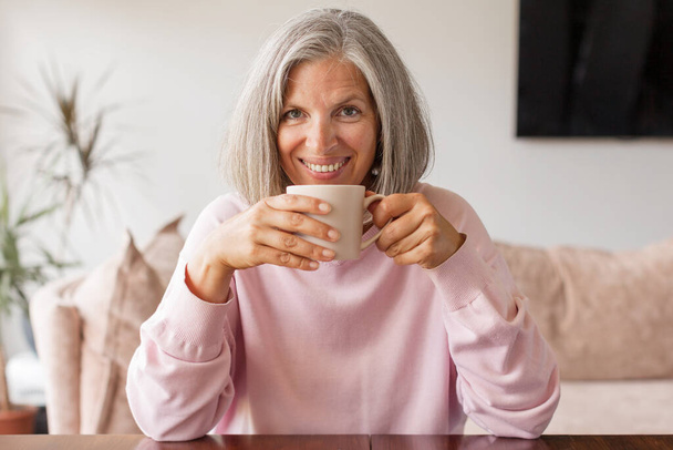 Mutlu, rahat, olgun, yetişkin, gri saçlı kadın evdeki kanepede kahve içiyor. Gülümseyen orta yaşlı kadın modern oturma odasındaki kanepede dinlenmekten zevk alıyor. - Fotoğraf, Görsel
