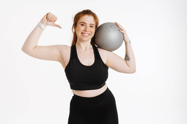 Sonriendo joven más tamaño fitness mujer en ropa deportiva de pie sobre fondo blanco ejercicio con una pelota de fitness - Foto, Imagen