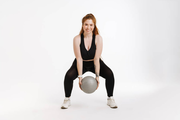 Sorridente giovane plus size fitness donna in abbigliamento sportivo in piedi su sfondo bianco esercizio con una palla fitness - Foto, immagini